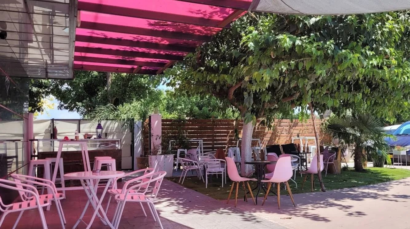 The terrace at Comvos Café in Amaliada: pink chairs and tables under a pink pergola and leafy trees.